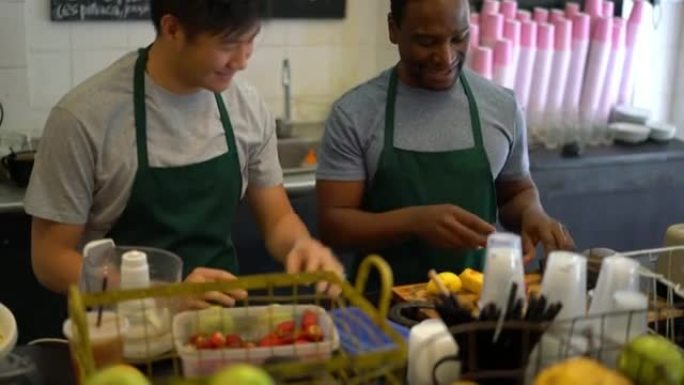 在果汁吧工作的黑人和亚洲推销员作为一个团队，一个选择水果，另一个切碎并将其添加到搅拌机中