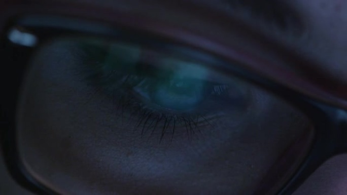 戴着眼镜的女人眼睛的特写镜头在黑暗中凝视着工作中的电脑屏幕。