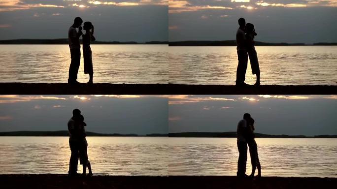日落时恋人的剪影。年轻美丽的夫妇在湖岸拥抱