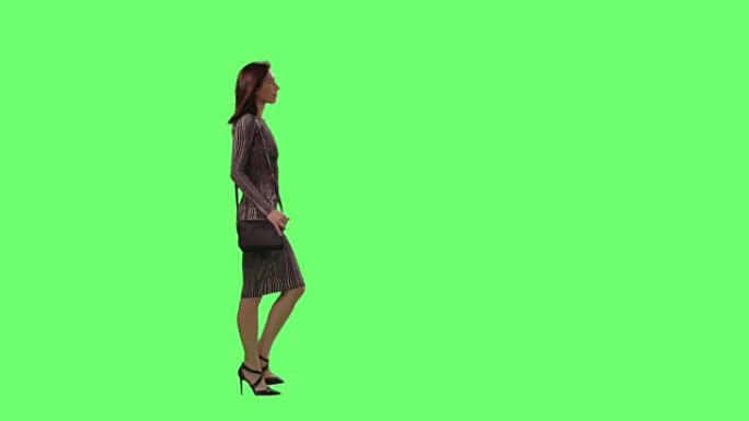 迷人的黑发女性穿着紧身连衣裙，带着一个小钱包，优雅地走在背景的模拟绿色屏幕上。