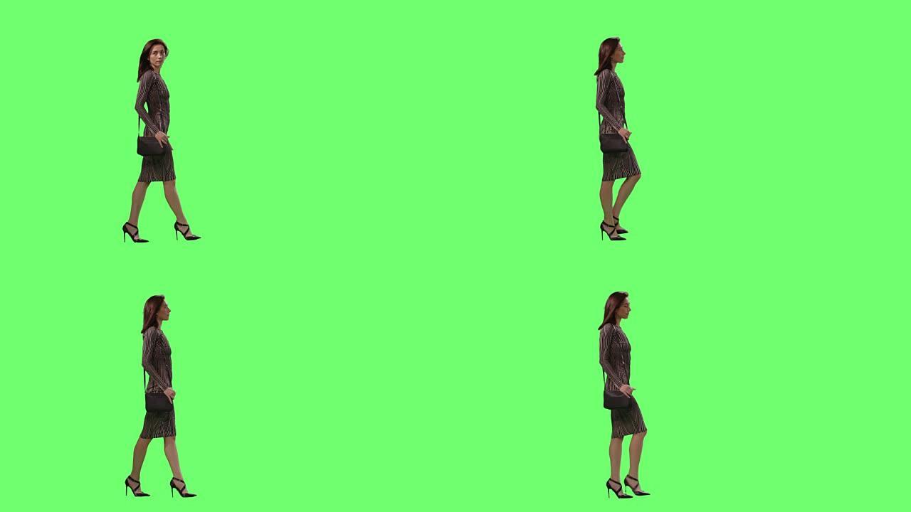迷人的黑发女性穿着紧身连衣裙，带着一个小钱包，优雅地走在背景的模拟绿色屏幕上。