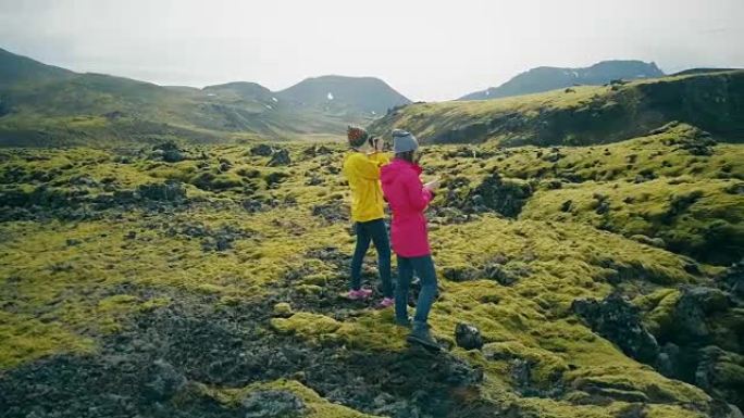 鸟瞰图，两名女游客在冰岛的熔岩上行走并拍摄照片。女孩使用智能手机和相机