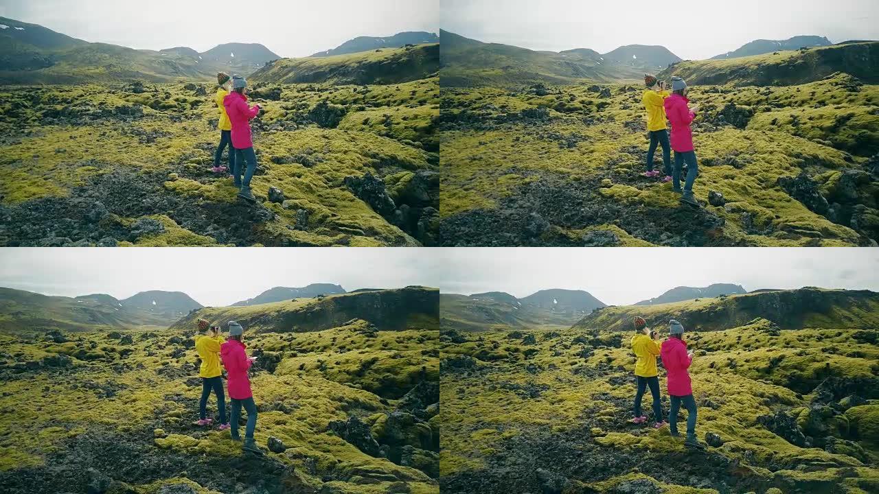鸟瞰图，两名女游客在冰岛的熔岩上行走并拍摄照片。女孩使用智能手机和相机