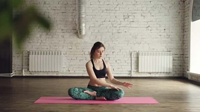 穿着时髦运动服装的年轻女士正坐在瑜伽馆内的垫子上做身体扭曲。放松个人实践理念。