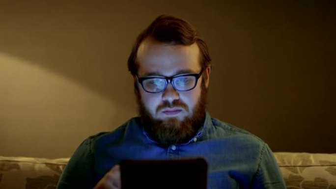 一名男子坐在客厅沙发上，使用平板电脑拍摄的肖像。落地灯打开。
