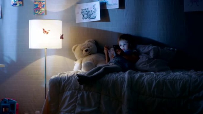 可爱的小女孩躺在床上，在智能手机上看动画片。她的落地灯亮着。