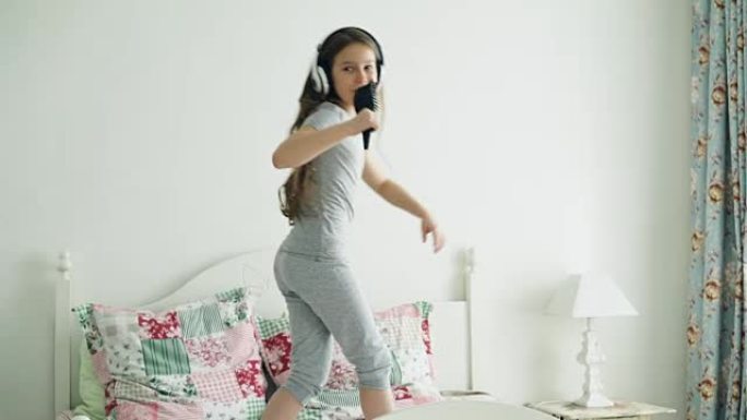 有趣可爱的小女孩戴着无线耳机，用梳子跳舞，在舒适的卧室里的床上玩得开心