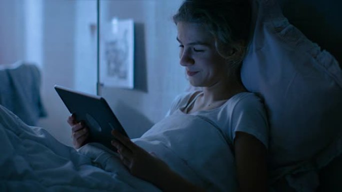 年轻的少女晚上躺在床上，用平板电脑。看电视节目，微笑。