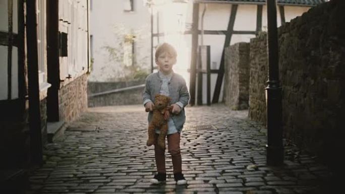 可爱的欧洲男孩在街上唱歌。男孩子抱着一只泰迪熊。才能表达。旧铺砌的城镇道路。4K