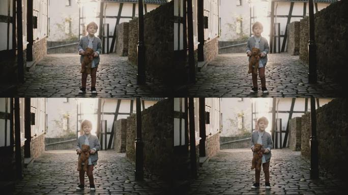可爱的欧洲男孩在街上唱歌。男孩子抱着一只泰迪熊。才能表达。旧铺砌的城镇道路。4K
