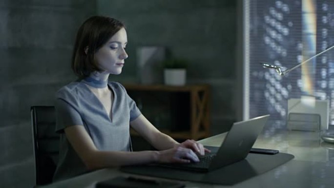 成功的年轻女子坐在办公桌前的私人办公室里，在笔记本电脑上工作。她的办公室以极简主义风格完成，很暗，窗