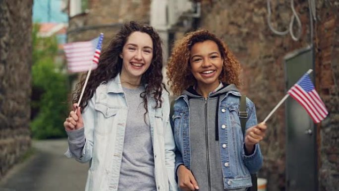 两个穿着休闲服装的漂亮女孩挥舞着美国国旗，笑着看着相机的慢动作肖像。友谊、旅游和幸福的人的概念。
