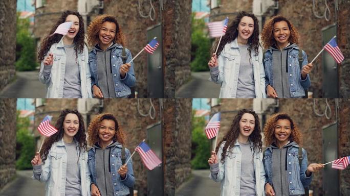 两个穿着休闲服装的漂亮女孩挥舞着美国国旗，笑着看着相机的慢动作肖像。友谊、旅游和幸福的人的概念。