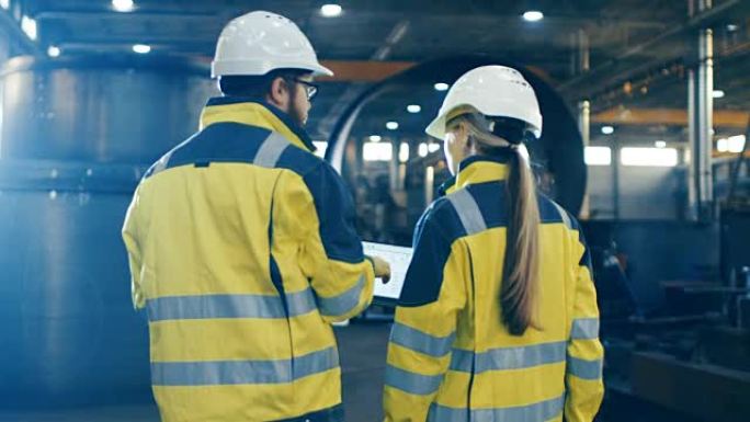 男女工业工程师在穿过重工业制造工厂时使用笔记本电脑并进行讨论。他们戴着安全帽和安全外套。背景大金属制