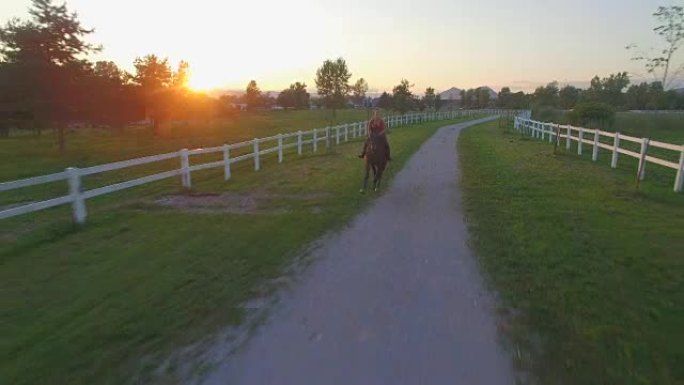 空中: 日落时分，年轻女孩在郊区小镇的田野里骑马
