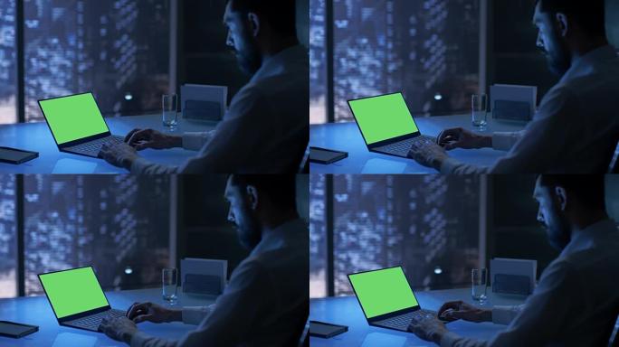 深夜商人在他的私人办公室里用一台带有绿色模拟屏幕的笔记本电脑工作，可以看到大城市的窗户。