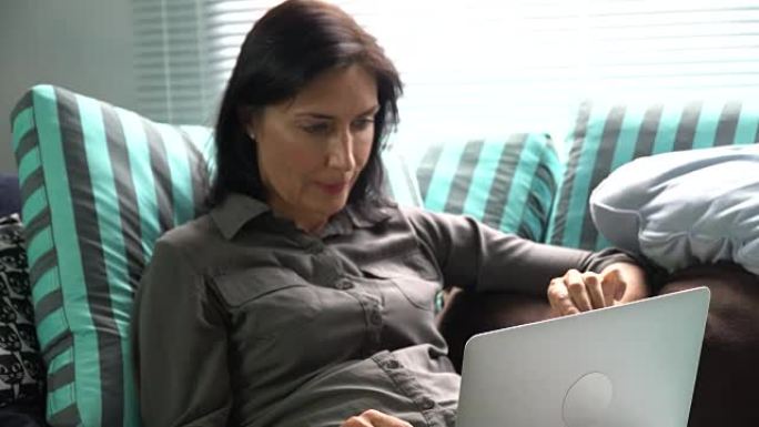 漂亮的女人坐在沙发上，家里有笔记本电脑。打字，浏览互联网