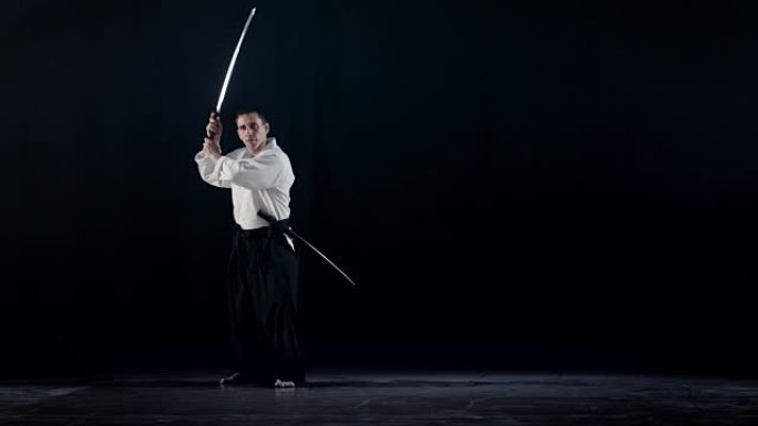 穿着传统的武士Hakama服装的合气道大师将他的日本刀从刀鞘中取出并摆动。他在聚光灯下，黑暗包围着他