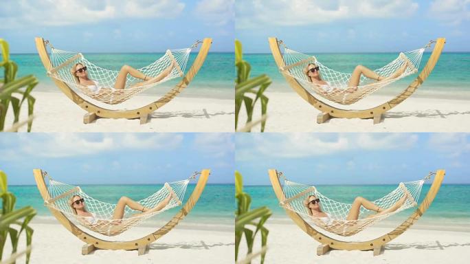 田园诗般的场景。美丽的女人躺在海滩的吊床上晒日光浴。有白色沙滩和海蓝宝石水的蔚蓝海滩。阳光照在驱魔的