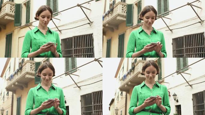 迷人的年轻黑发女人穿着浅色夏装在欧洲小镇的街道上。她正在看着手机并使用它。
