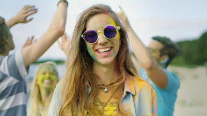 一个戴着墨镜的美丽年轻女孩的特写肖像站在庆祝胡里节的人群中。人们在她背上扔五颜六色的粉末。她的脸上和