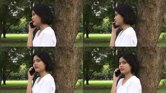 高清格式: 快乐的年轻女子坐在秋公园的树下在手机上聊天。