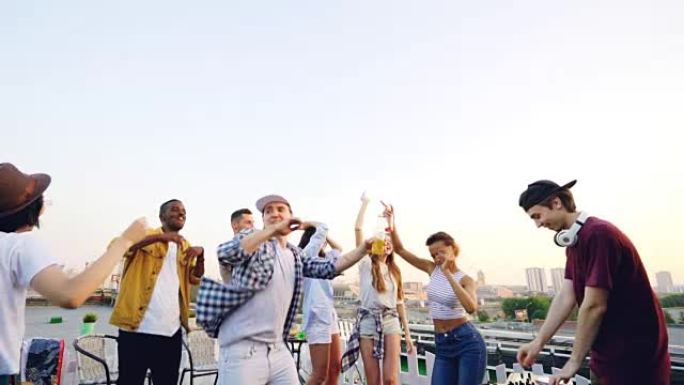 夏季，男性DJ在屋顶派对上使用设备时，年轻人的慢动作跳舞和大笑。娱乐、青春和音乐概念。