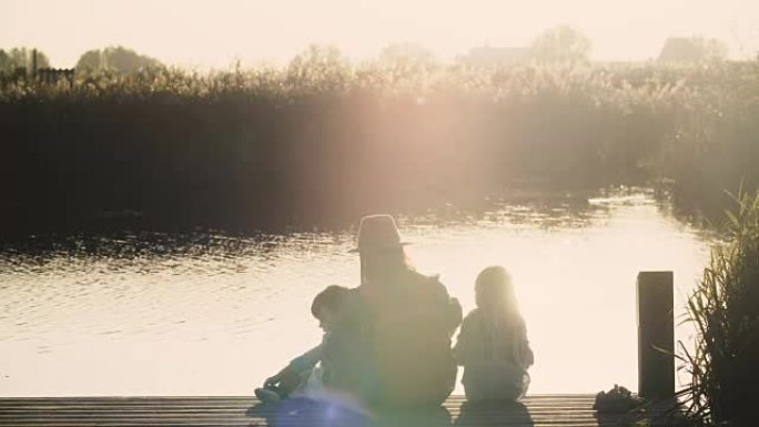 母亲和两个孩子在日出时坐在湖码头上。和平与和谐。家庭交往时间。惊人的温暖镜头耀斑。4K