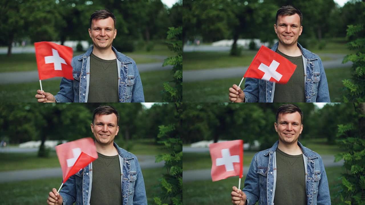 瑞士体育迷微笑着的慢动作肖像，挥舞着瑞士的旗帜，看着背景为绿树和草坪的相机。青年与民族主义概念。