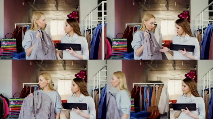 年轻的企业主站在她的服装店时正在使用平板电脑。她的助手带着衣服来征求意见。女商人正在向她展示放置它的