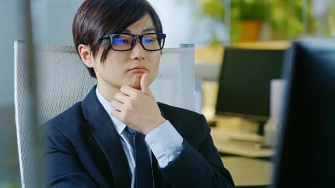 日本商人穿着西装和眼镜，坐在办公桌前，在个人电脑上工作的肖像。他在考虑问题解决方案。