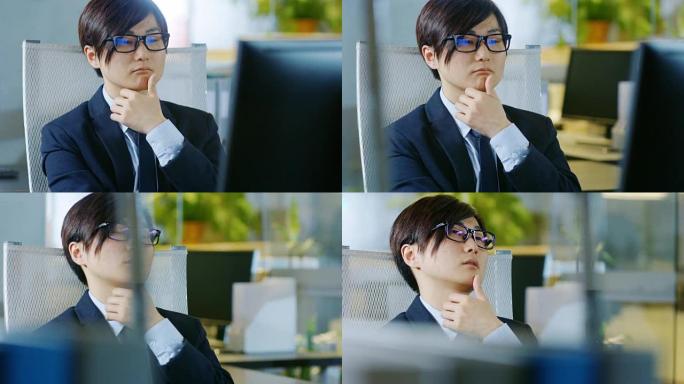 日本商人穿着西装和眼镜，坐在办公桌前，在个人电脑上工作的肖像。他在考虑问题解决方案。
