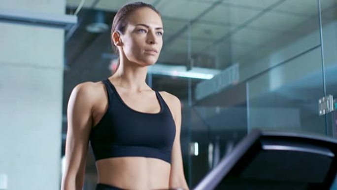 美丽的女人运动员穿着运动胸罩，在跑步机上行走，使用触摸屏选择最佳速度。在背景现代健身房/健身俱乐部。