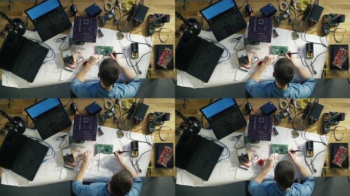 年轻的it技术人员焊接电路板的俯视图。他的桌子上摆满了各种计算机零件，主板，笔记本电脑，电缆。早晨的