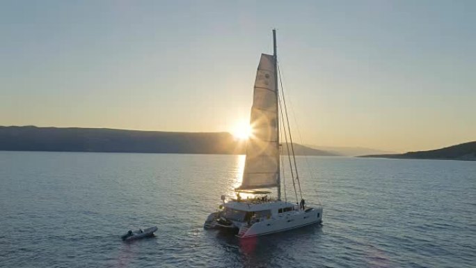 一艘帆船双体船的空中拍摄，帆船在平静的海面上航行，太阳升起在山后。人们在甲板上放松。