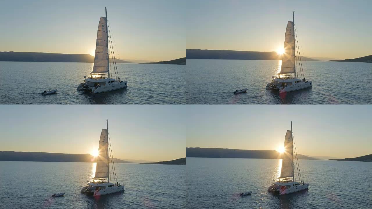 一艘帆船双体船的空中拍摄，帆船在平静的海面上航行，太阳升起在山后。人们在甲板上放松。