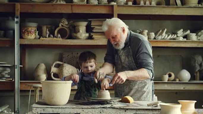 可爱的男孩在他有爱心的祖父教书并帮助他，并提供建议时，正在在投掷轮上模制粘土。背景中有许多陶瓷锅的架