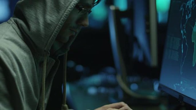 头罩中的男性黑客在黑暗的办公室里的显示屏上显示地图和数据的计算机上工作。