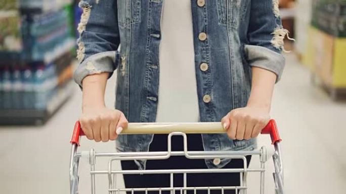 特写镜头女性双手推着超市里的购物车，货架上摆放着食物和饮料。购买和选择产品和人的概念。