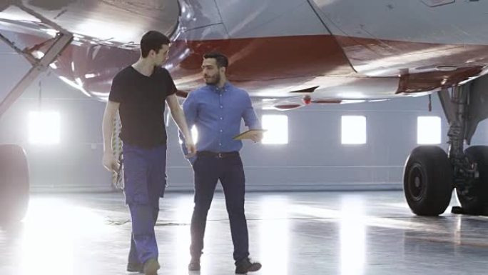 在机库，飞机维修工程师向飞机技术员展示平板电脑上的技术数据。他们与干净的全新飞机同行。