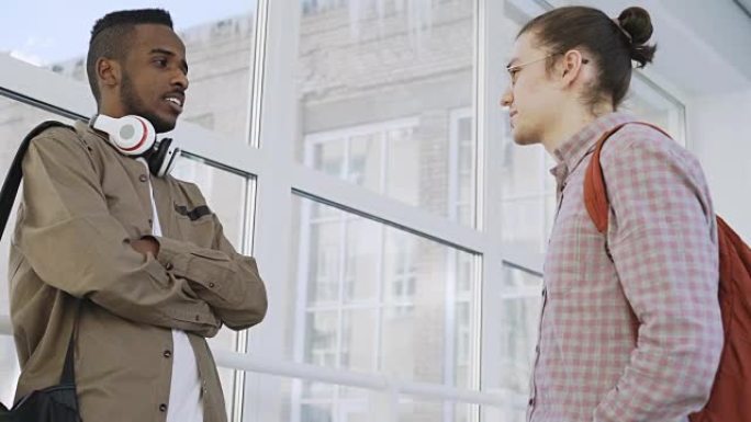 两个年轻的帅哥正站在玻璃般的大学走廊里，以积极的方式交流并微笑着。非洲学生正在向高加索人解释一些事情