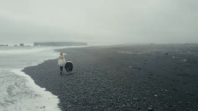 在冰岛黑色火山海滩上散步的年轻夫妇的鸟瞰图。lopapeysa的男人和女人在海上扔石头