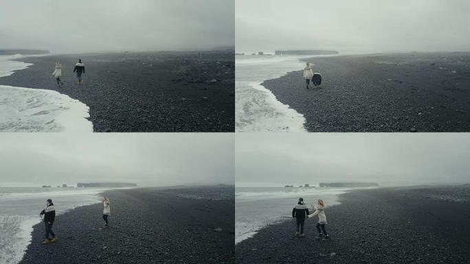 在冰岛黑色火山海滩上散步的年轻夫妇的鸟瞰图。lopapeysa的男人和女人在海上扔石头