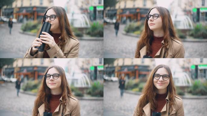 戴着眼镜的年轻漂亮法国学生的肖像站在街上，戴着帽子喝茶，对着镜头微笑