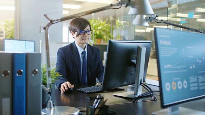 在办公室里，东亚商人在台式个人电脑上工作。他同事的电脑显示信息图