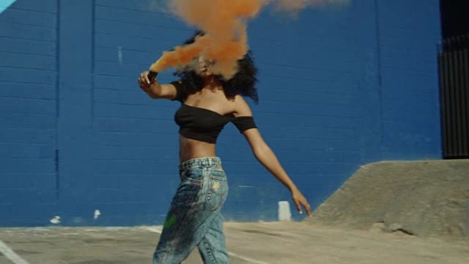 美丽的年轻女子手持五颜六色的烟雾弹对着蓝墙在外面跳舞