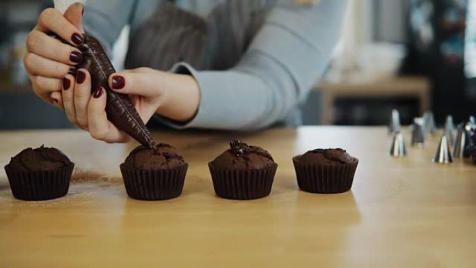 女性手的特写镜头将糕点袋中的奶油放在巧克力纸杯蛋糕上。年轻女子烹饪松饼