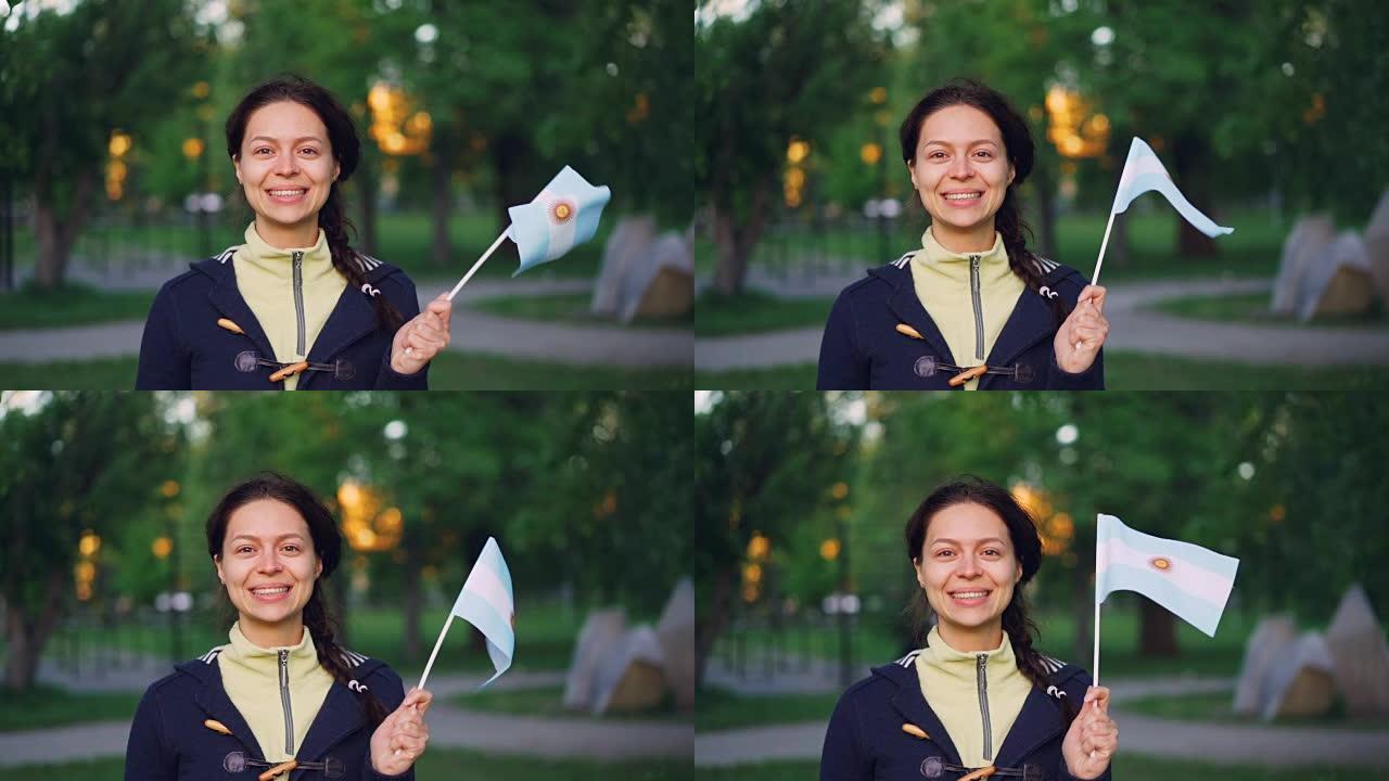 微笑的阿根廷年轻女士的慢动作肖像挥舞着阿根廷国旗站在绿色城市公园外面，看着相机。