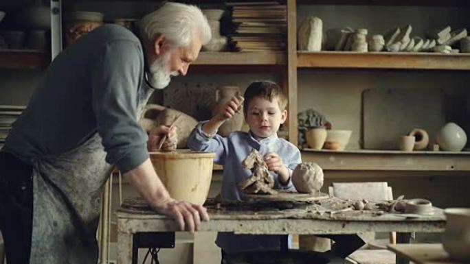 可爱的孩子在他祖父的工作室里玩粘土和波特的工具制作陶瓷玩具。灰发的大胡子男人正带着爱和关怀和谈话看着