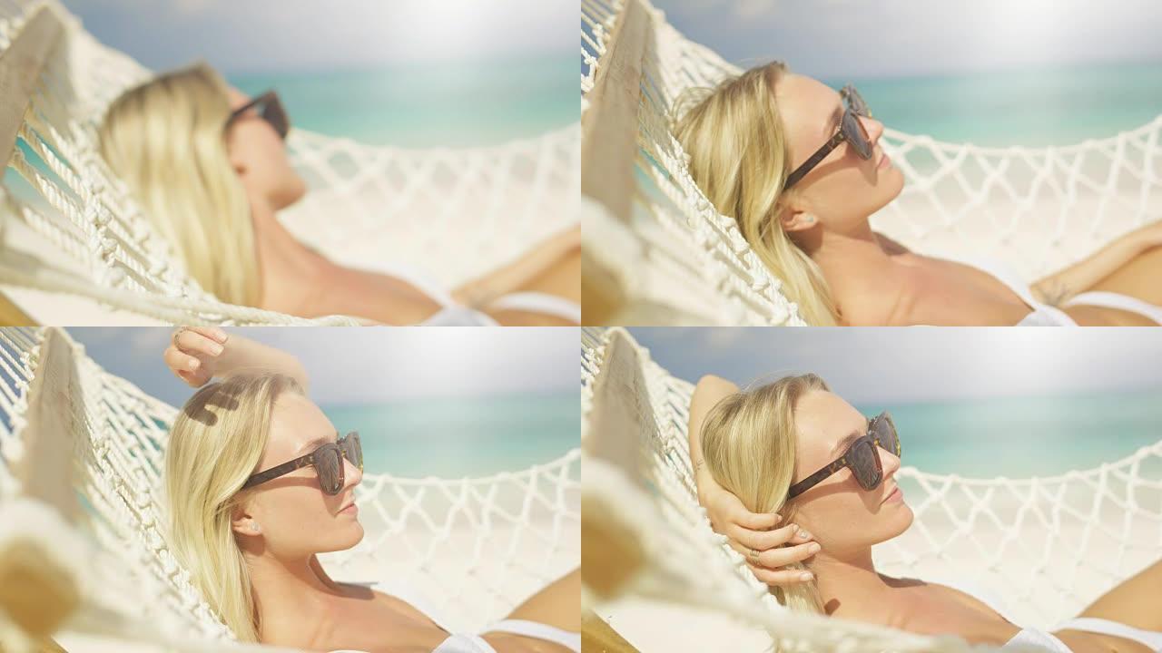 田园诗般的场景。一个戴着太阳镜的金发美女的特写镜头躺在海滩的吊床上晒太阳。有白色沙滩和海蓝宝石水的蔚